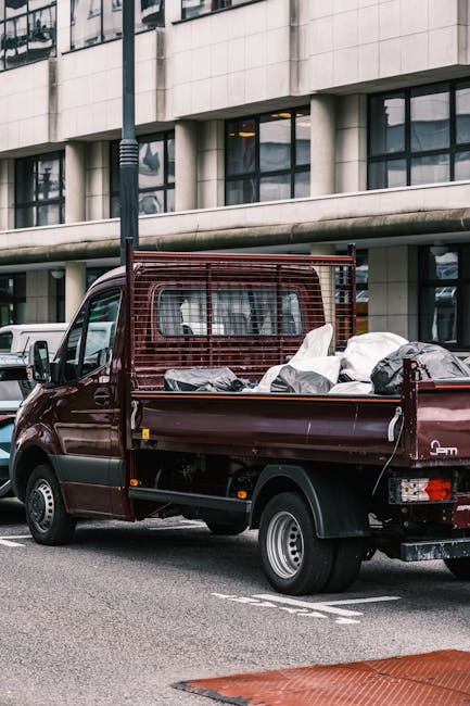 A small burgundy flatbed truck parked alongside an urban street, filled with a variety of waste materials including dark black and grey plastic bags, cardboard boxes, and other assorted rubbish. The t